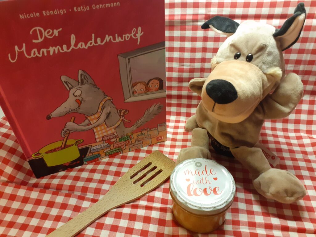 Handpuppe Wolf mit Kochlöffel und dem Buch "Der Marmeladenwolf" auf karierter Tischdecke, daneben ein Marmeladenglas
